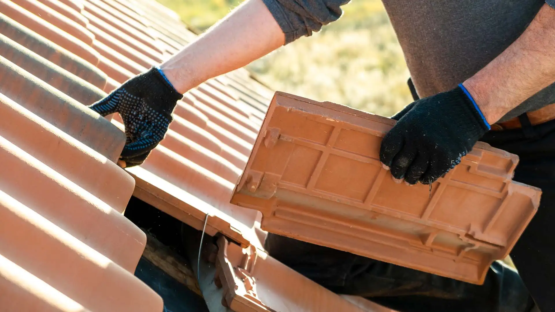 Gros plan d'un couvreur utilisant des gants de protection pour remplacer une tuile cassée sur une toiture en terre cuite.